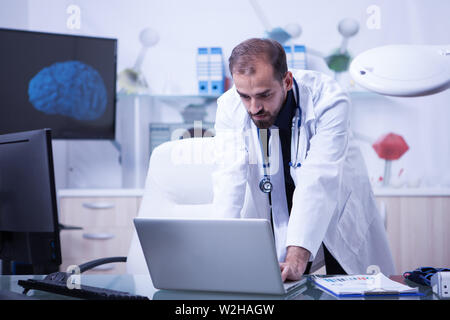 Arzt auf seinem Laptop und Kontrolle der Behandlung seiner Patienten. Attraktive Arzt mit einem weißen Mantel. Stockfoto