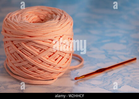 Hobbys und kreative Aktivitäten Konzept - eine Spule von großen Gestrickte Garne aus Pfirsich Farbe mit orangefarbenen glänzenden Haken auf der Oberfläche der blauen Marmor. Stockfoto