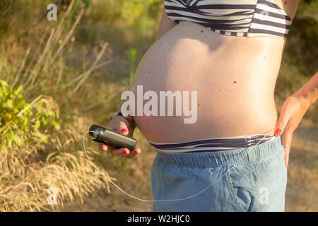 Eine diabetische schwangere Frau mit einer Insulinpumpe in einem Wald. Sie trägt einen blauen Rock und einem gestreiften Bikini Top. Stockfoto