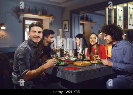 Ein junger Mann mit einem Glas ist lächelnd vor dem Hintergrund von Freunden in einem Restaurant. Stockfoto