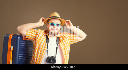 Fat lustiger Mann mit Bart in einem Sommer Shirt in einen Hut mit einem Koffer lächelnd geht auf grauem Hintergrund. Das ursprüngliche Konzept des World, Deutsch, Freizeit, Reisen in Stockfoto