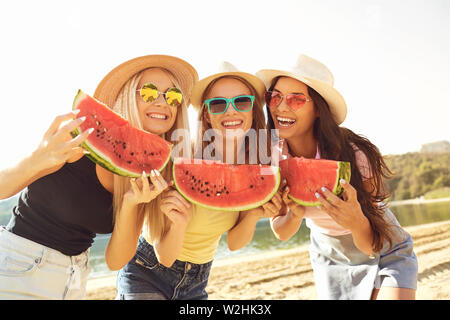 Schöne glückliche Frauen in Sonnenbrillen und Hüte, die auf sandigen Küste mit Scheiben Wassermelone lächelnd an Kamera im Sonnenlicht Stockfoto