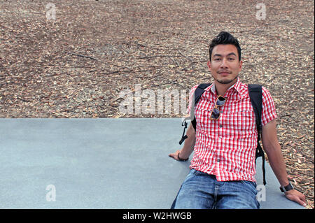 Junger Mann mit dunklem Haar und Bart trägt ein kariertes Hemd und einen Rucksack sitzt auf einem Betonblock Stockfoto