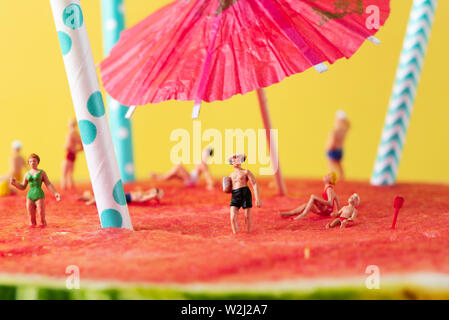 Einige miniatur Menschen tragen Badeanzug entspannen auf einen erfrischenden reife Wassermelone, mit weißen und blauen Trinkhalme und ein Papier Regenschirm klemmt Ich Stockfoto