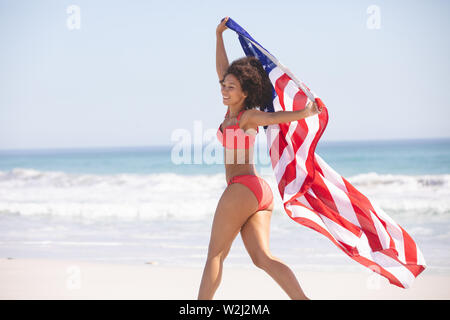 Frau im Bikini mit amerikanischer Flagge zu Fuß am Strand Stockfoto