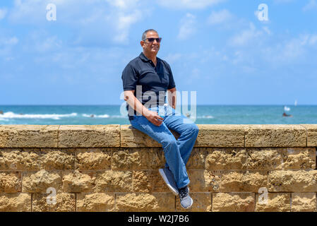 Lächelnd trendy Touristische älterer Mann sitzt von alten Ziegel historische Stein. Portrait von Stattlichen alter Mode Senior mit schwarzen T-Shirt und Jeans auf Bl Stockfoto