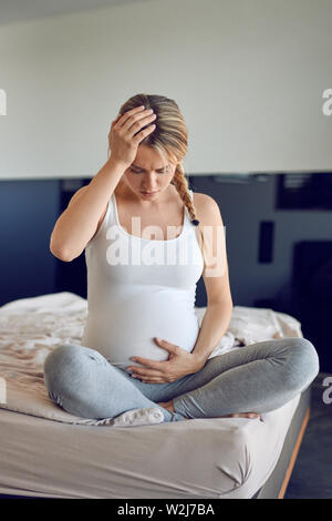 Stark gestörten schwangere junge Frau sitzt im Schneidersitz auf einem Bett mit ihrem aufgeblähten Bauch und hielt den Kopf gesenkt, mit einem ernsten Ex Stockfoto