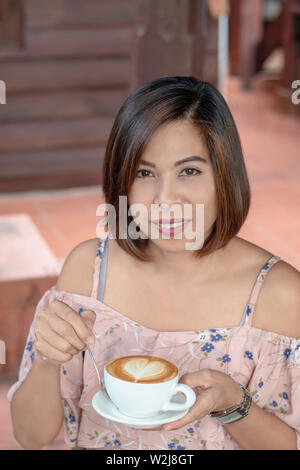 Hand asiatische Frau mit heißem Kaffee Espresso gekrönt mit einem Herzförmigen Milch in weißem Glas. Stockfoto