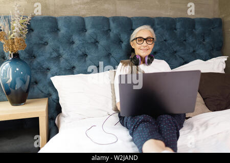 Alte Frau mit dem grauen Haar in Brillen Tragen von Kopfhörern Festlegung auf dem Bett mit Laptop Stockfoto