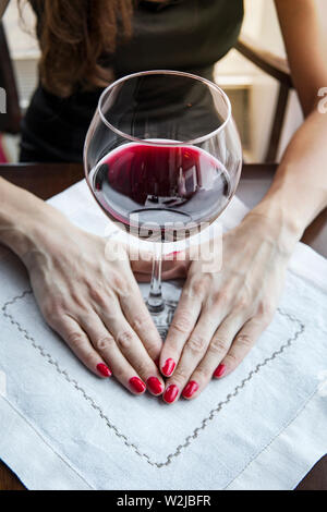 Die Hand einer Frau mit einem roten Nagel hält einen Becher mit Rotwein, ein großes Glas Rotwein auf einer weißen Serviette auf dem Tisch im Restaurant. Stockfoto