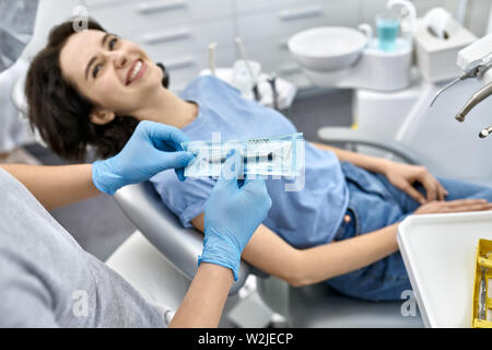 Lächelnde Mädchen liegt auf einem Stuhl in einer Zahnarztpraxis. Zahnarzt in einem grauen Uniform und schützende blaue Handschuhe hält eine versiegelte zahnmedizinische Bur. Horizontale Foto mit Stockfoto