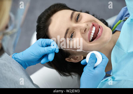 Happy girl in Bib eines Patienten in einer Zahnarztpraxis. Sie bewegt ihre Zunge über ihre Zähne. Zahnarzt in blau Latex Handschuhe bereitet sich auf ihre Zähne flossing mit Stockfoto