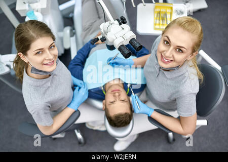 Schöner Mann in Bib des Patienten in einer Zahnarztpraxis. Zwei lächelnde Zahnärzte in einem grauen Uniform und blau Latex Handschuhe sind neben ihm. Ansicht von oben Hori Stockfoto