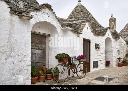 Toller alter Trulli auf dem bewölkten Himmel Hintergrund in Alberobello Stadt in Italien. Es gibt viele bunte Blumen in Töpfe, vintage Fahrradverleih in der Nähe von Stockfoto