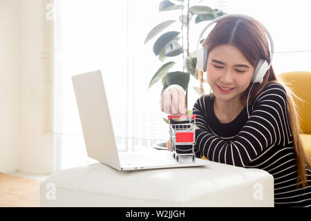 Junge asiatische Frau im Zimmer sitzen und mit Notebook Computer für Online Shopping und Musik mit Kopfhörer zu Hause Stockfoto