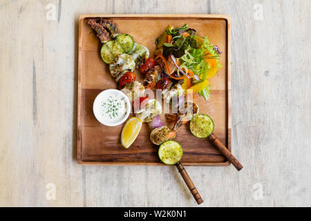 Nahaufnahme der gemischte gegrillte Meeresfrüchte Spieße mit gesunden Salat und Sauce, Garnelen, Jakobsmuscheln, Calamari, Lachs, Möhre, Salat, in ein Holzbrett platter und Stockfoto