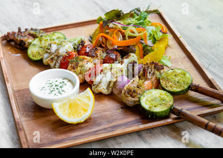 Nahaufnahme der gemischte gegrillte Meeresfrüchte Spieße mit gesunden Salat und Sauce, Garnelen, Jakobsmuscheln, Calamari, Lachs, Möhre, Salat, in ein Holzbrett platter und Stockfoto