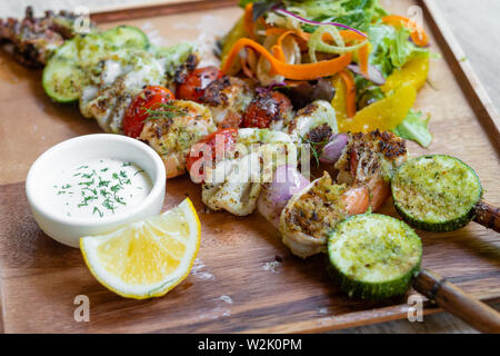 Nahaufnahme der gemischte gegrillte Meeresfrüchte Spieße mit gesunden Salat und Sauce, Garnelen, Jakobsmuscheln, Calamari, Lachs, Möhre, Salat, in ein Holzbrett platter und Stockfoto