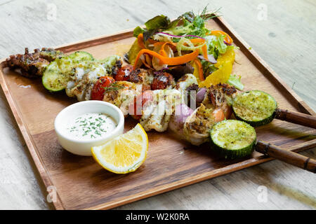 Nahaufnahme der gemischte gegrillte Meeresfrüchte Spieße mit gesunden Salat und Sauce, Garnelen, Jakobsmuscheln, Calamari, Lachs, Möhre, Salat, in ein Holzbrett platter und Stockfoto