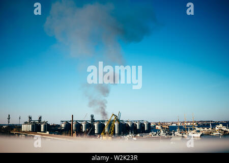 Blick aus der Ferne der industriellen Hafen und Fabrik mit schwarzen Rauch, der die Wogen in blauer Himmel es umweltschädliche. Rauch von Feuer an industrielle Anlage. Stockfoto