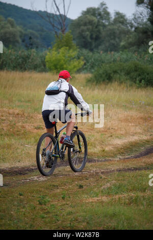 Charkow, Ukraine - Juli 06, 2019: Ansicht von hinten von einem Mann mit dem Fahrrad auf einem Feldweg in einem Feld in der Nähe des Waldes Stockfoto
