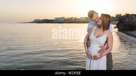 Schwangere Frau umarmen und küssen mit ihrem Mann am Strand bei Sonnenuntergang Stockfoto