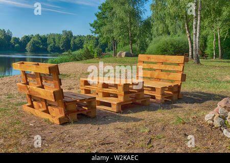 Rustikalen Holztisch und Bänken mit Paletten am Ufer eines ruhigen See von Bäumen und viel Grün im Sommer Sonne umgeben Stockfoto