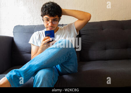 Spanischer junger Mann mit klaren braunen Haut mit Brille mit verwirrt Smartphone, während auf einer Couch Stockfoto