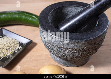 Mörser und Stößel aus Stein auf einem Küchentisch mit Kartoffel, Hanf samen und Gurke Stockfoto