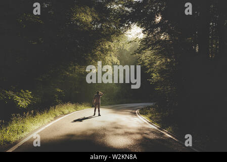 Wanderer zu Fuß auf der Straße in einem schattigen Wald. Morgen Sonnenstrahlen durch die Bäume auf die Straße kommen. Stockfoto