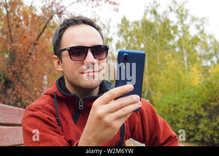Europäische Kerl sitzt auf einer Bank in einer Stadt Herbst Park. Ein Mann ist ein Lächeln am Telefon. Konzept der Freude und Vergnügen, Freude und Lachen. Humor Stockfoto
