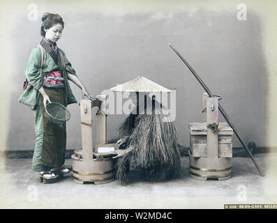 [1880s Japan - Japanische Tofu Anbieter] - ein Tofu (Tofu) Hersteller tragen eines Mino (Stroh regenmantel) und einem sugegasa Konische hat Schichten einen Block von Tofu mit einem Messer. Eine Frau im Kimono und traditionelle Frisur hält einen Korb. Eine sehr ökologische Weise Tofu zu verkaufen! 19 Vintage albumen Foto. Stockfoto