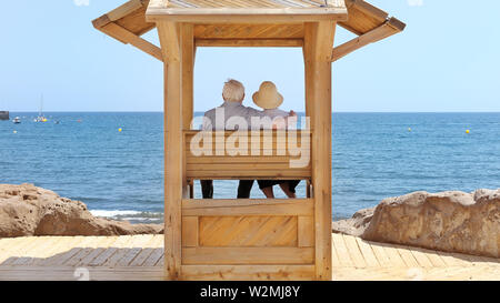 Senior Paar, Frau mit Hut und Mann mit weissen silbernes Haar sitzen unter einem hölzernen Baldachin auf der Promenade und Blick auf das Meer in El Medano Stockfoto