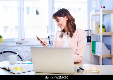 Junge Frau mit Smartphone sitzt von Schreibtisch bei der Arbeit Stockfoto