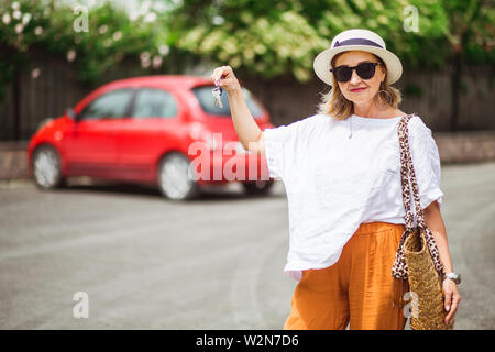 Schöne Frau mit Schlüssel zum neuen Auto gekauft und lächelnd an der Kamera in Sonnenbrille und Hut im Sommer. Stockfoto