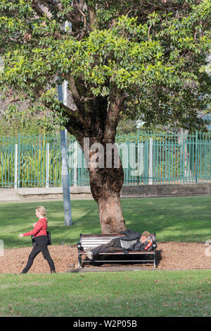 Eine Frau, die an einem obdachlosen älteren Mann vorbeigeht, der auf einer Parkbank in einem Stadtpark in Sydney in Australien schläft Stockfoto