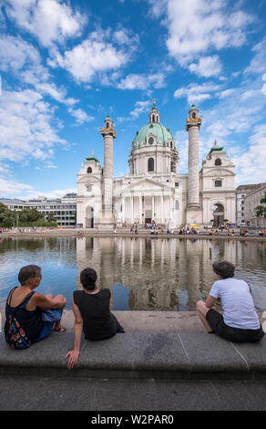 Menschen neben dem Teich an der Karlskirche (St Charles Kirche), eine Barocke römisch-katholische Kirche und die örtlichen Sehenswürdigkeiten, Wien, Österreich sitzen. Stockfoto