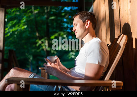 Mann sitzt auf Schaukelstuhl auf der Veranda des Hauses im Morgen Blockhaus cottage Kaffee Trinken aus dem Becher Stockfoto