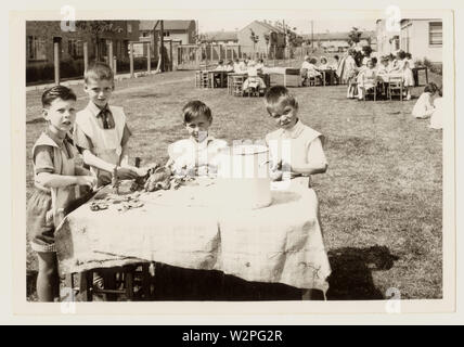 1960er Jahre Foto von Grundschulkindern, die Schürzen oder Lätzchen tragen, mit Spielteig auf Tischen spielen, die auf den Schulspielfeldern draußen, im Freien, möglicherweise Leicestershire, Großbritannien, aufgestellt sind Stockfoto