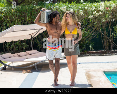 Interracial Paare, schwarzer Mann und kaukasische Frau zu Fuß mit einer Kassette Radio am Pool Stockfoto