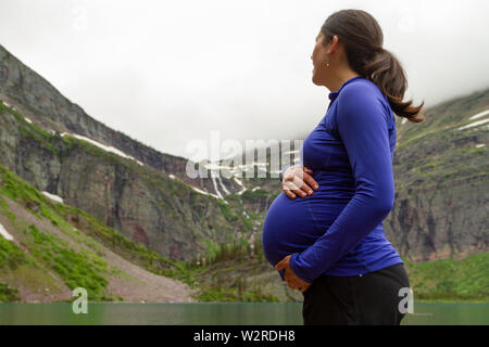 Frau in den späten dritten Trimester, nähert sich dem Ende ihrer Schwangerschaft, liebevoll mit ihrem Bauch am Ufer des Sees, Grinnell Glacier National Park. Stockfoto