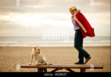 Mitte - erwachsener Mann mit Mantel und Krone beim Stehen auf Tisch am Strand mit seinem Hund. Stockfoto