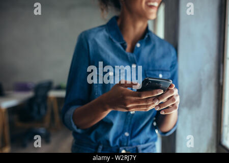 Frau in Casuals im Büro durch ein Fenster mit Telefon in der Hand. Geschäftsfrau mit Handy in der Hand. Stockfoto