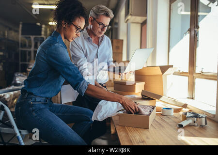 Online Store Partner Vorbereitung ein Paket für den Versand. Frau Verpackung das Produkt in Box mit Menschen arbeiten am Laptop. Stockfoto