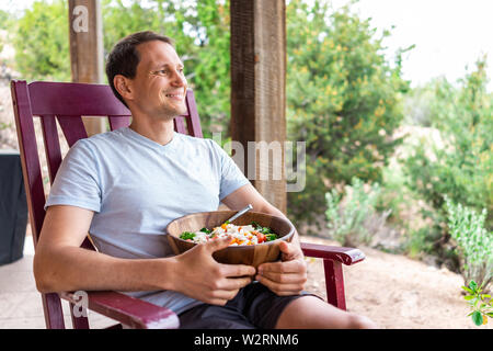 Glücklich Mann mit frischem Salat in Houten mit Salat sitzen auf Schaukelstuhl in Garten Terrasse Stockfoto