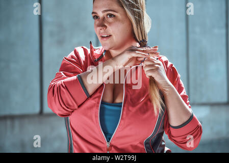 Bereit, mein Bestes zu tun. Cute plus size Frau in Sportkleidung Anpassung ihre langen Haare und suchen weg, während im Freien stehen. Übergewichtige Frau. Sportkonzept Stockfoto