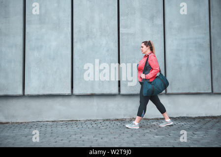 Das Training ist vorbei. Rückansicht der Plus-Größe Frau in Sportkleidung tragen ihre Tasche und nach Hause gehen nach Übungen im Freien. Sportkonzept. Übergewichtige Frau Stockfoto