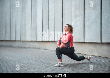Ich liebe Sport glücklich plus size Frau in Sportkleidung Stretching-Übungen und lächeln, während sie gegen die graue Wand stehen. Gesundes Leben. Sportkonzept. Abnehmen. Stockfoto