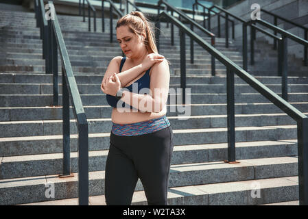Schmerzen nach dem Training. Müde Plus size Frau in Sport Kleidung Leiden von der Schulter Schmerzen beim Stehen auf Treppen. Sportverletzung. Gewicht zu verlieren. Stockfoto