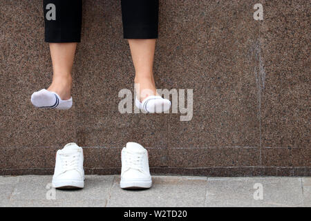 Müde Beine, Fußschmerz, barfuss Frau mit entfernten weißen Turnschuhen ruht auf einer Straße. Weibliche Beine in weißen Socken über Schuhen baumelt. Stockfoto